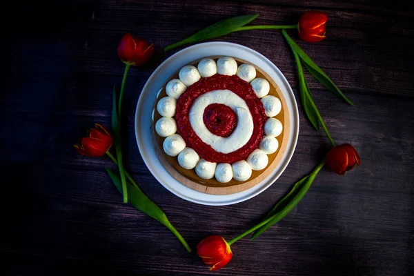 Медовый бисквит с украшением из белого воздушного крема и слоями красной малины стоит на белом блюде на темном деревянном фоне, тюльпаны лежат по кругу — стоковое фото