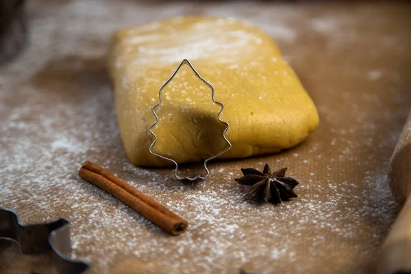 A cookie cutter in the form of a Christmas tree is leaning on the dough, cinnamon and cloves are lying next to it, flour, other cookie cutters and a rolling pin for dough are visible in the background