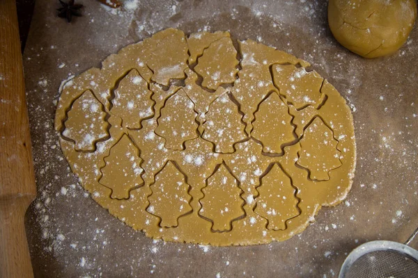 Dans la couche roulée de la pâte, de nombreux arbres de Noël sont découpés, qui sont marqués avec des contours, la pâte repose sur le parchemin de cuisson, à côté de lui se trouve un rouleau à pâtisserie, une passoire pour la farine, tout — Photo