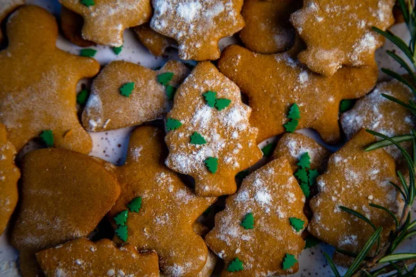 Kurabiyeler farklı şekillerde bir tabakta, ön planda bir Noel ağacı şeklinde bir kurabiye var, kar gibi pudra şekerle süslenmiş ve küçük yeşil Noel ağaçlarıyla süslenmiş.