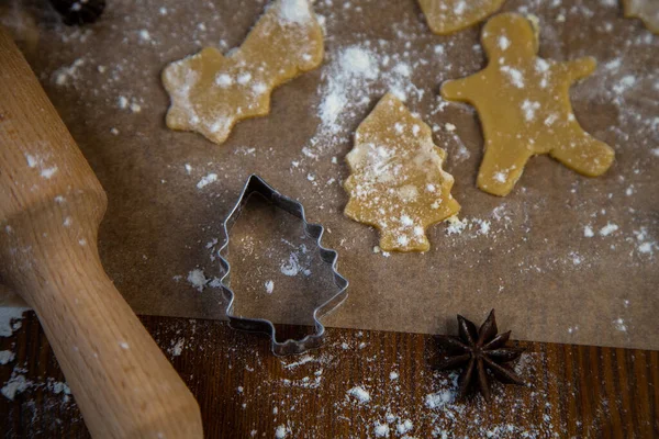 Le moule pour biscuits en forme d'arbres de Noël sur le parchemin couché à côté de lui sont coupés des morceaux de pâte sur le parchemin, à côté d'un rouleau à pâtisserie et toute la farine sur un fond sombre — Photo