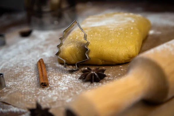 A cookie cutter in the form of a Christmas tree is leaning on the dough, cinnamon and cloves are lying next to it, flour, other cookie cutters and a rolling pin for dough are visible in the background