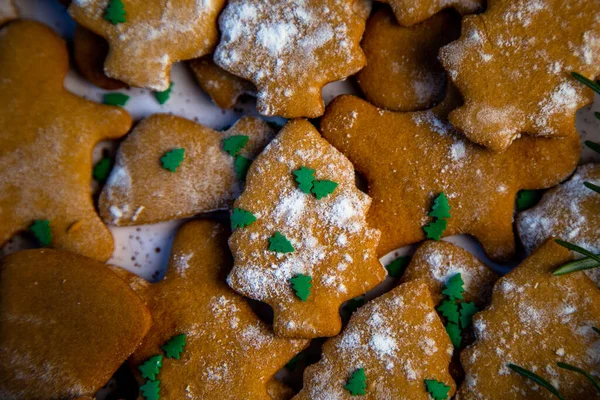 Τα cookies βρίσκονται σε ένα πιάτο διαφορετικών σχημάτων, σε πρώτο πλάνο υπάρχει ένα μπισκότο με τη μορφή χριστουγεννιάτικου δέντρου, διακοσμημένο με άχνη ζάχαρη όπως το χιόνι και μικρά πράσινα χριστουγεννιάτικα δέντρα ενάντια στο — Φωτογραφία Αρχείου