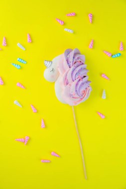 Çubukta beyaz, pembe-mor kuyruklu ve gümüş boynuzlu bir meringue tek boynuzlu atı, parlak sarı bir arka planda uzanır, etrafına renkli toz boynuzları saçılmıştır. dikey fotoğrafçılık