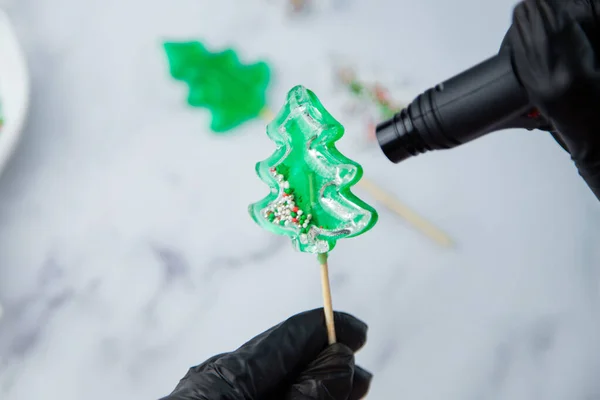 Una piruleta verde en forma de árbol de Navidad se sostiene en las manos de guantes negros y se quema en el fuego de un encendedor, en el proceso de hacer una piruleta. —  Fotos de Stock