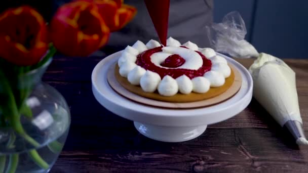 Dark sponge cakes stand on a white stand, they are decorated with white cream, a pastry chef from a pastry bag applies raspberry jam between the brown cream. — Stockvideo