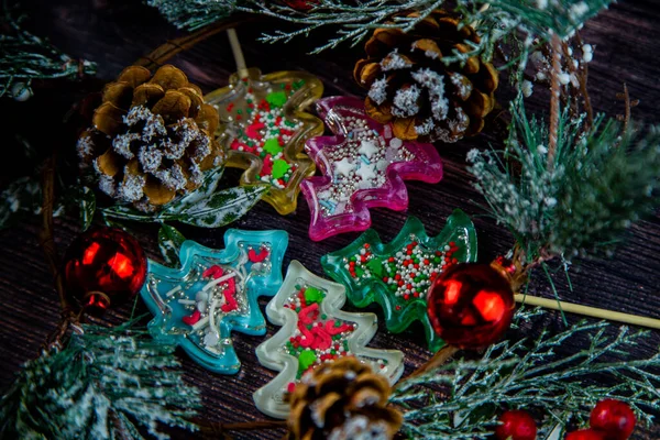A lot of Christmas trees of lollipops of different colors lie in a circle on a wooden table surrounded by branches of a Christmas tree, cones and Christmas toys. — Foto Stock