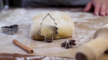 Un içindeki koyu bir parşömenin üzerinde bir parça hamur var, üzerine noel ağacı şeklinde bir kurabiye kesicisi eğiliyor, bir oklava, diğer kalıplar, tarçın ve karanfiller onun yanında, beyaz