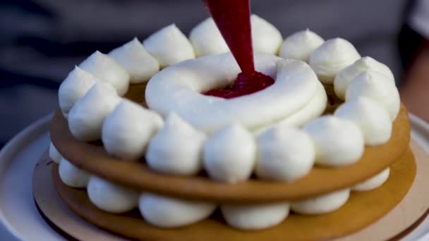 Dark sponge cakes stand on a white stand, they are decorated with white cream, a pastry chef from a pastry bag applies raspberry jam between the brown cream. — 图库视频影像