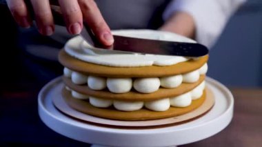 Aşçı pastanın üst krema tabakasını pürüzsüz hale getirmek için demir bir spatula kullanır ve pastayı kürsüye kaydırır. Video yakın planda ve siyah bir anahtarla çalışıyor.