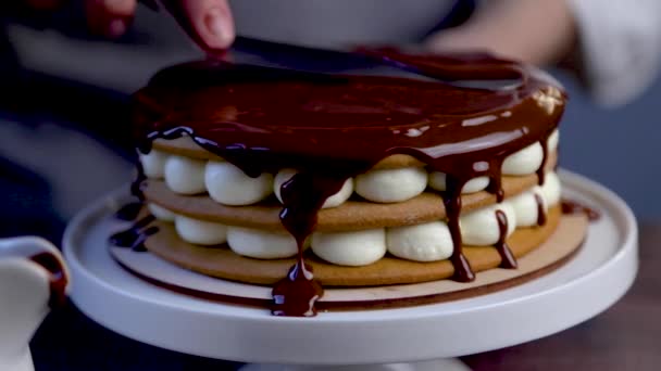 Торт, що складається з трьох шарів, тортів і білого крему, стоїть на підставці, прикрашений білим кремом і розтопленим шоколадом зверху, а кухар розподіляє шоколад по поверхні за допомогою заліза — стокове відео