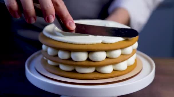 Повар использует железную лопатку, чтобы сгладить верхний кремовый слой торта, прокручивая торт на подставке. Видео крупным планом и выполнено в темном ключе — стоковое видео