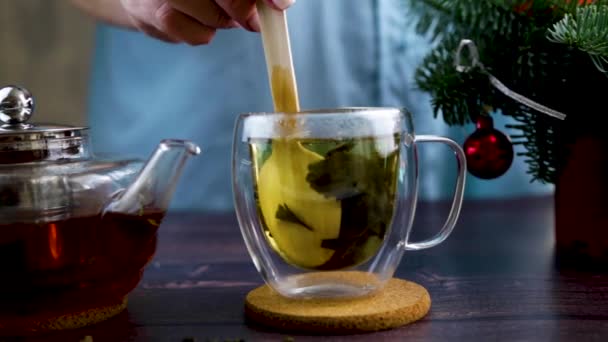 Женская рука берет деревянную ложку и перемешивает чай с чайными листьями внутри стеклянной кружки. Рядом есть елка и прозрачный чайник с чаем, видео в темном ключе — стоковое видео