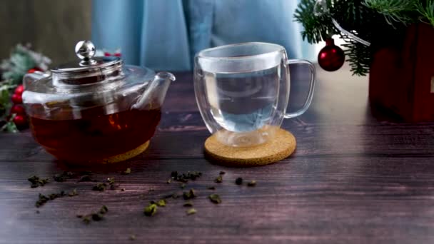 Une tasse en verre est sur la table, de l'eau bouillante est versée dans la tasse, dans laquelle une main abaisse une cuillère en bois avec du thé, des bulles proviennent des pétales de thé, une théière avec du thé est à côté, les feuilles de thé sont — Video