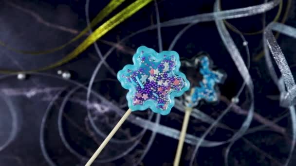 A lollipop in the form of a snowflake - rattles of blue color are shaking from side to side, inside the balls are moving, against a background of dark marble and ribbons. — Stock Video