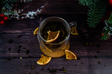 Şeffaf bir fincan çay koyu bir masada, ahşap bir standın üzerinde duruyor, kurumuş portakal dilimleri fincanda yüzüyor, yanında narenciye dilimleri uzanıyor, çay dağınık, siyah bir anahtardaki fotoğraf.