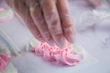 Beyaz ponponları ve kenarları olan pembe kremalı şapkaların kenarında beyaz toplar serpiştiriliyor, parşömen arka planına karşı.