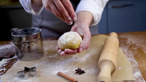 Kucharz ciasta trzyma ciasto w ręku, posypuje mąką drugą ręką, film jest w ciemnym kluczu, z wałkiem, pergaminem, uchwytem na mąkę i ciasteczkami w tle. — Wideo stockowe