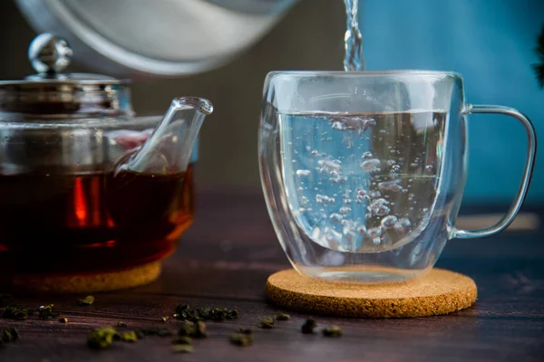 Hot water is poured into a transparent mug from a teapot, bubbles are in the water, a teapot and tea leaves are in the background