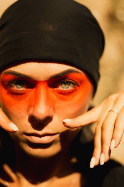 Portrait of a young beautiful woman with makeup and face art in American tribal or Middle Eastern style. Ethnic image, red eyes and black headscarf. Close-up.