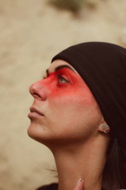 Portrait of a young beautiful woman with makeup and face art in American tribal or Middle Eastern style. Ethnic image, red eyes and black headscarf. Close-up.