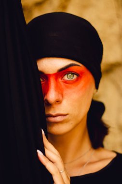Portrait of a young beautiful woman with makeup and face art in American tribal or Middle Eastern style. Ethnic image, red eyes and black headscarf. Close-up.