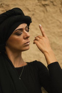 Makyajlı, beyaz yüzlü, kabile ya da orta doğu tarzında siyah tesettüre bürünmüş güzel bir kadının portresi. Kumlu bir duvarın arka planında etnik görüntü.