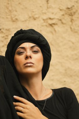 Makyajlı, beyaz yüzlü, kabile ya da orta doğu tarzında siyah tesettüre bürünmüş güzel bir kadının portresi. Kumlu bir duvarın arka planında etnik görüntü.
