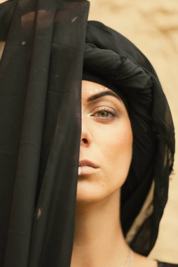Makyajlı, beyaz yüzlü, kabile ya da orta doğu tarzında siyah tesettüre bürünmüş güzel bir kadının portresi. Kumlu bir duvarın arka planında etnik görüntü.