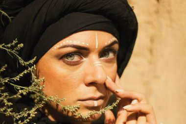 Makyajlı, beyaz yüzlü, kabile ya da orta doğu tarzında siyah tesettüre bürünmüş güzel bir kadının portresi. Kumlu bir duvarın arka planında etnik görüntü.