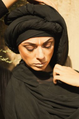 Makyajlı, beyaz yüzlü, kabile ya da orta doğu tarzında siyah tesettüre bürünmüş güzel bir kadının portresi. Kumlu bir duvarın arka planında etnik görüntü.
