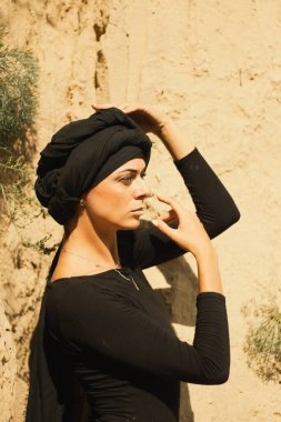 Makyajlı, beyaz yüzlü, kabile ya da orta doğu tarzında siyah tesettüre bürünmüş güzel bir kadının portresi. Kumlu bir duvarın arka planında etnik görüntü.