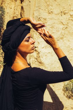 Makyajlı, beyaz yüzlü, kabile ya da orta doğu tarzında siyah tesettüre bürünmüş güzel bir kadının portresi. Kumlu bir duvarın arka planında etnik görüntü.