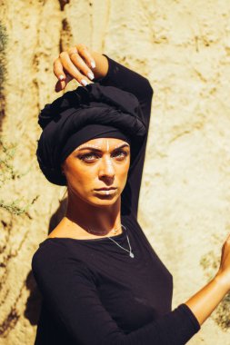 Makyajlı, beyaz yüzlü, kabile ya da orta doğu tarzında siyah tesettüre bürünmüş güzel bir kadının portresi. Kumlu bir duvarın arka planında etnik görüntü.