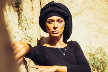 Makyajlı, beyaz yüzlü, kabile ya da orta doğu tarzında siyah tesettüre bürünmüş güzel bir kadının portresi. Kumlu bir duvarın arka planında etnik görüntü.