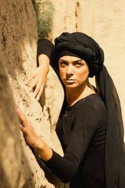 Makyajlı, beyaz yüzlü, kabile ya da orta doğu tarzında siyah tesettüre bürünmüş güzel bir kadının portresi. Kumlu bir duvarın arka planında etnik görüntü.