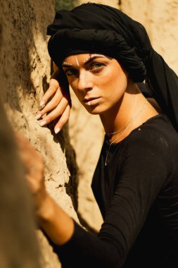 Makyajlı, beyaz yüzlü, kabile ya da orta doğu tarzında siyah tesettüre bürünmüş güzel bir kadının portresi. Kumlu bir duvarın arka planında etnik görüntü.
