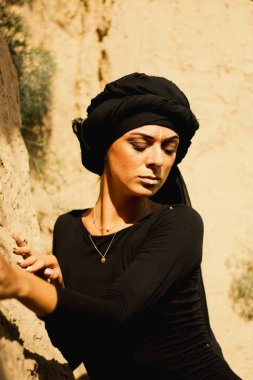 Makyajlı, beyaz yüzlü, kabile ya da orta doğu tarzında siyah tesettüre bürünmüş güzel bir kadının portresi. Kumlu bir duvarın arka planında etnik görüntü.