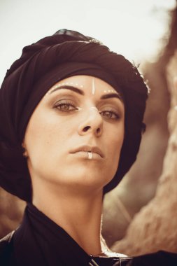 Makyajlı, beyaz yüzlü, kabile ya da orta doğu tarzında siyah tesettüre bürünmüş güzel bir kadının portresi. Kumlu bir duvarın arka planında etnik görüntü.