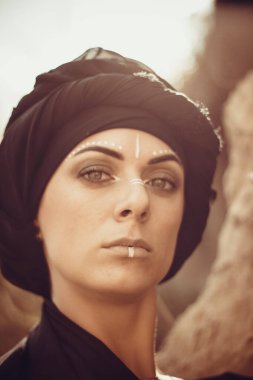 Makyajlı, beyaz yüzlü, kabile ya da orta doğu tarzında siyah tesettüre bürünmüş güzel bir kadının portresi. Kumlu bir duvarın arka planında etnik görüntü.