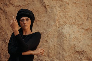 Siyah tesettüre bürünmüş, dans eden, kabile ya da ortadoğu tarzında makyaj ve beyaz yüz sanat süsü olan güzel bir kadının portresi. Kumlu duvarın arka planında etnik resim