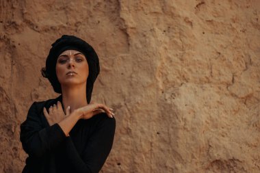 Siyah tesettüre bürünmüş, dans eden, kabile ya da ortadoğu tarzında makyaj ve beyaz yüz sanat süsü olan güzel bir kadının portresi. Kumlu duvarın arka planında etnik resim