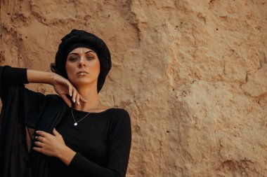 Siyah tesettüre bürünmüş, dans eden, kabile ya da ortadoğu tarzında makyaj ve beyaz yüz sanat süsü olan güzel bir kadının portresi. Kumlu duvarın arka planında etnik resim