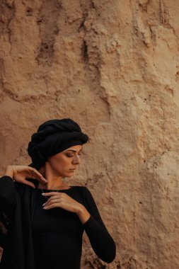 Siyah tesettüre bürünmüş, dans eden, kabile ya da ortadoğu tarzında makyaj ve beyaz yüz sanat süsü olan güzel bir kadının portresi. Kumlu duvarın arka planında etnik resim
