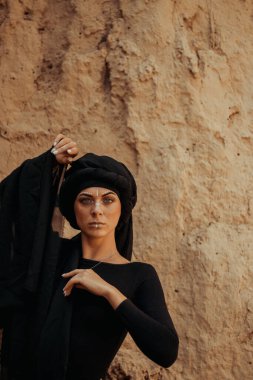 Siyah tesettüre bürünmüş, dans eden, kabile ya da ortadoğu tarzında makyaj ve beyaz yüz sanat süsü olan güzel bir kadının portresi. Kumlu duvarın arka planında etnik resim