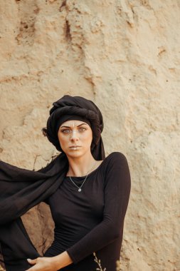 Siyah tesettüre bürünmüş, dans eden, kabile ya da ortadoğu tarzında makyaj ve beyaz yüz sanat süsü olan güzel bir kadının portresi. Kumlu duvarın arka planında etnik resim