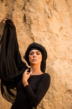 Siyah tesettüre bürünmüş, dans eden, kabile ya da ortadoğu tarzında makyaj ve beyaz yüz sanat süsü olan güzel bir kadının portresi. Kumlu duvarın arka planında etnik resim