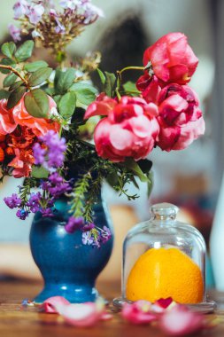 Bir buket parlak gül çiçeği, masasında mavi bir vazoda sardunyalar, bir portakal, yapraklar. Doğal ışık, yakın çekim, güzel arkaplan, dokular.