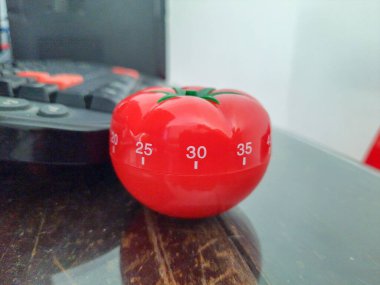 Pomodoro mutfak zamanlayıcısı ahşap bir masanın üzerinde duran kırmızı domates şeklinde ve etrafında renkli nesneler var.