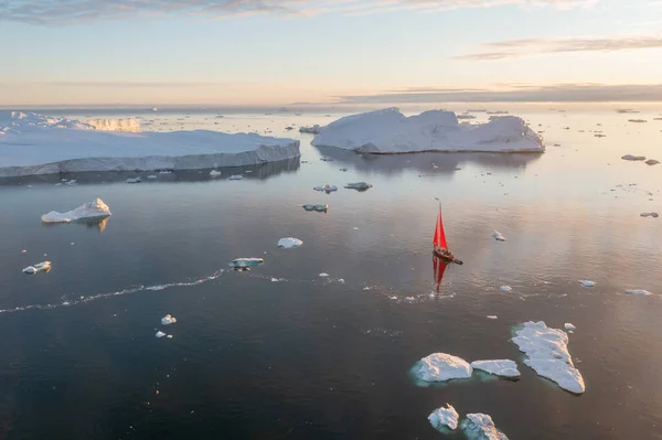 Sail boat with red sails cruising among ice bergs during dusk in front of a full moon. Disko Bay, Greenland. Midnight sun, romantic view. Climate change and global warming